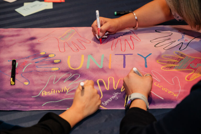 Global Refuge staff members collaborate on an activity in a ballroom - close up of hands writing on a poster that reads "Unity"